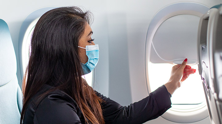 A masked passenger on a plane adjusting the window shade.