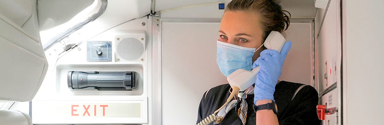 A flight attendant wearing a mask and gloves making an announcement to passengers.