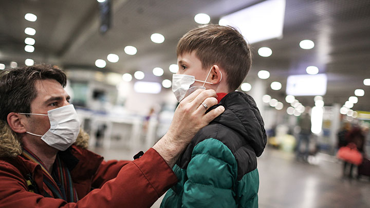 A father protecting his young son's health by adjusting his mask in a busy international airport.